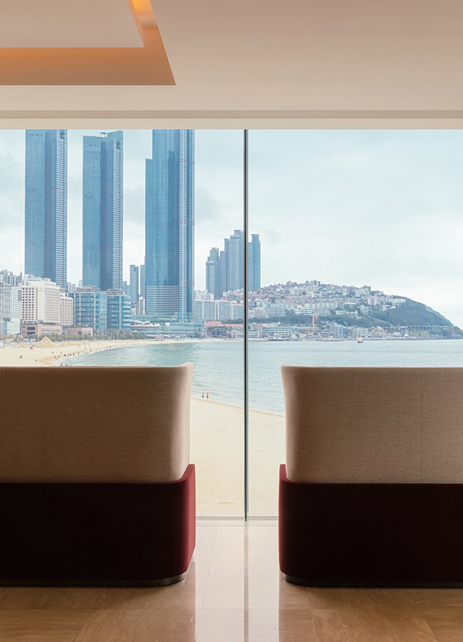 Lounge seating by a floor-to-ceiling window faces Haeundae Beach and the Marine City skyline.