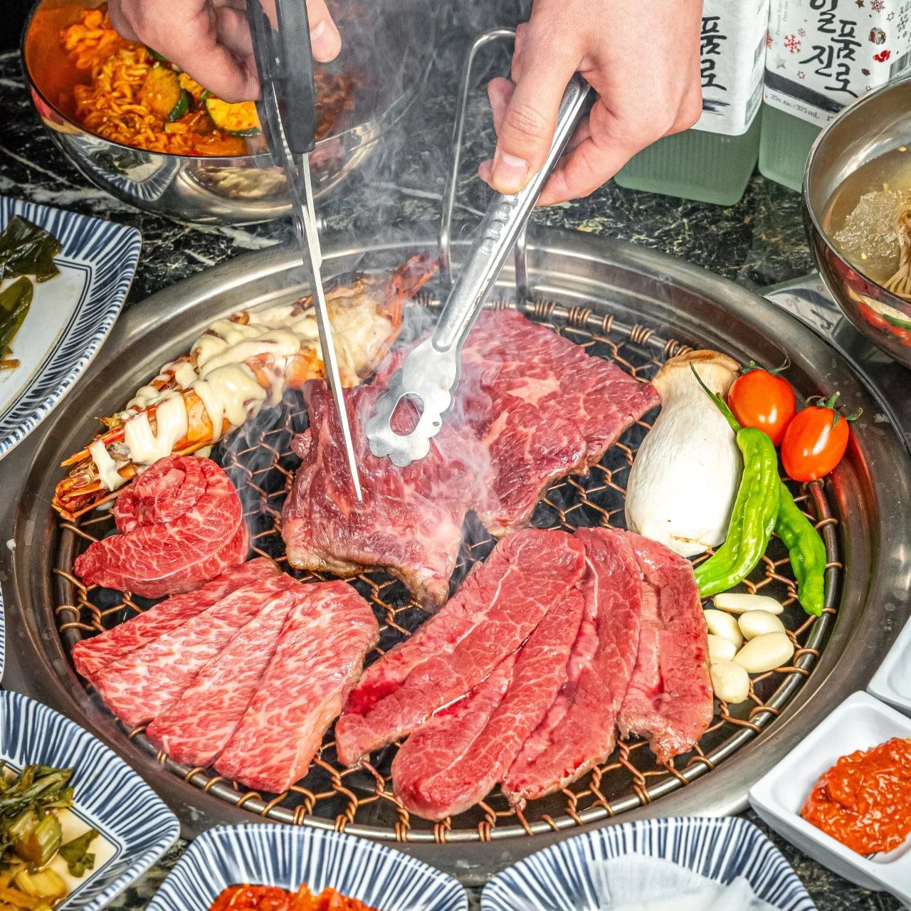 Assorted cuts of beef cooking with vegetables and a prawn on a circular tabletop grill.