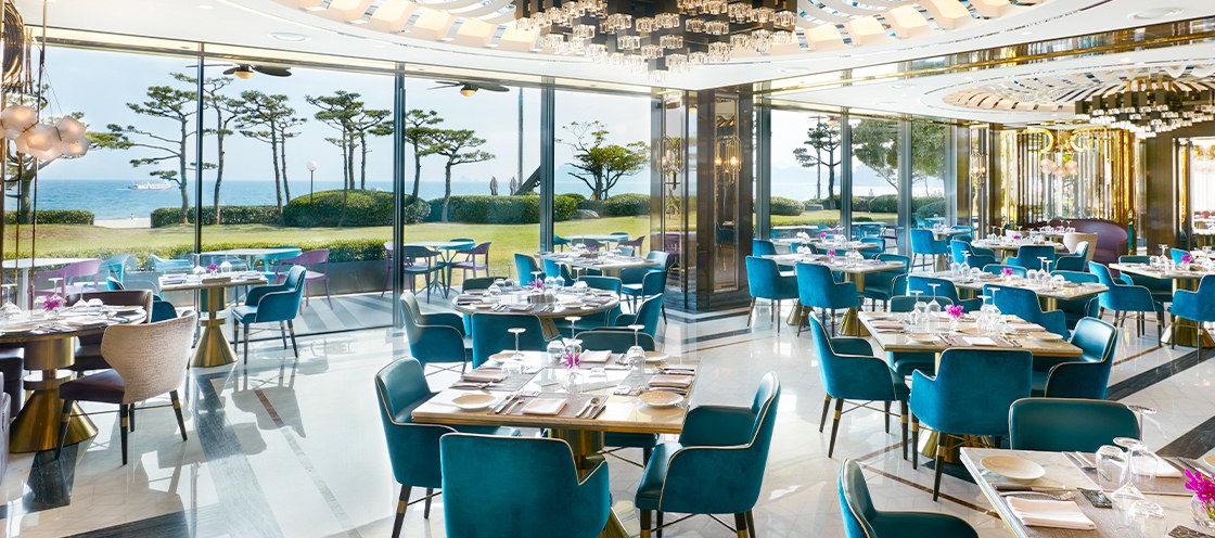 On the Plate’s dining room with turquoise chairs, set tables, and garden and sea views through full-height windows. The official image file is stored under a March 2021 path and remained displayed on the official page on September 20, 2026.