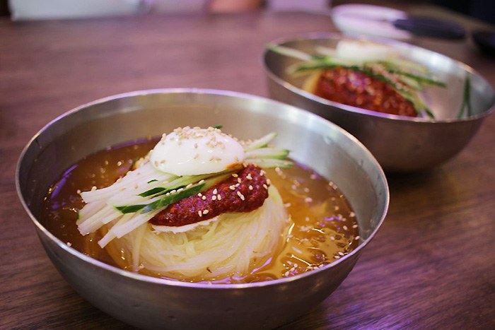 Two bowls of milmyeon topped with red seasoning, cucumber, egg and sesame seeds.